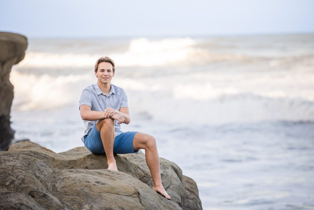A young man sits on a boulder at the beach with waves crashing behind him. 
