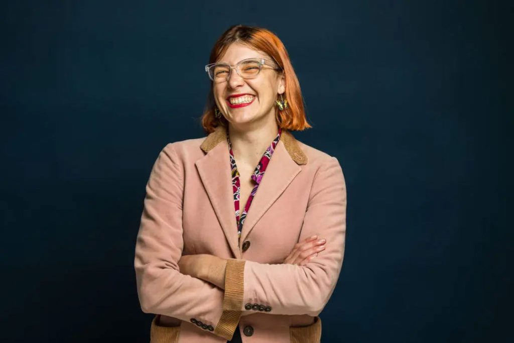 Woman with a pink coat and red printed innerwear with her amrs crossed while smiling brightly for her headshot