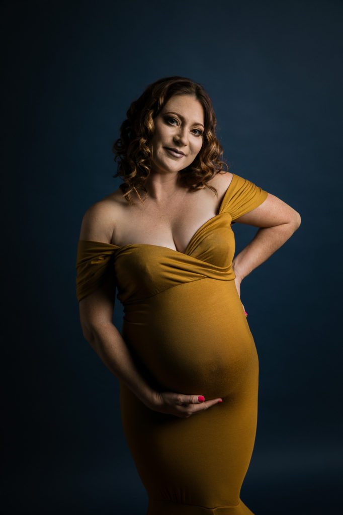 Woman wearing an ochre-colored off shoulder dress while clutching her pregnant belly