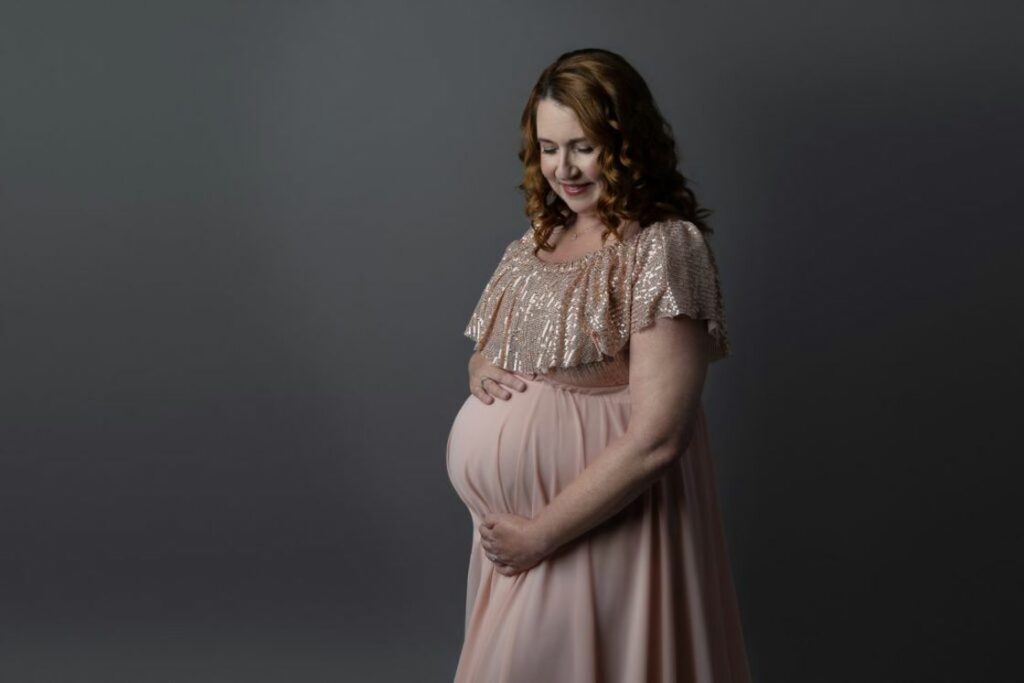 Woman wearing a long peach dress with a shimmery top, cupping her pregnant belly for her maternity shoot