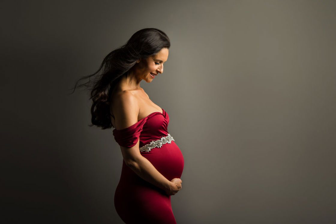 Woman wearing a red off shoulder dress holding her pregnant belly for her maternity shoot