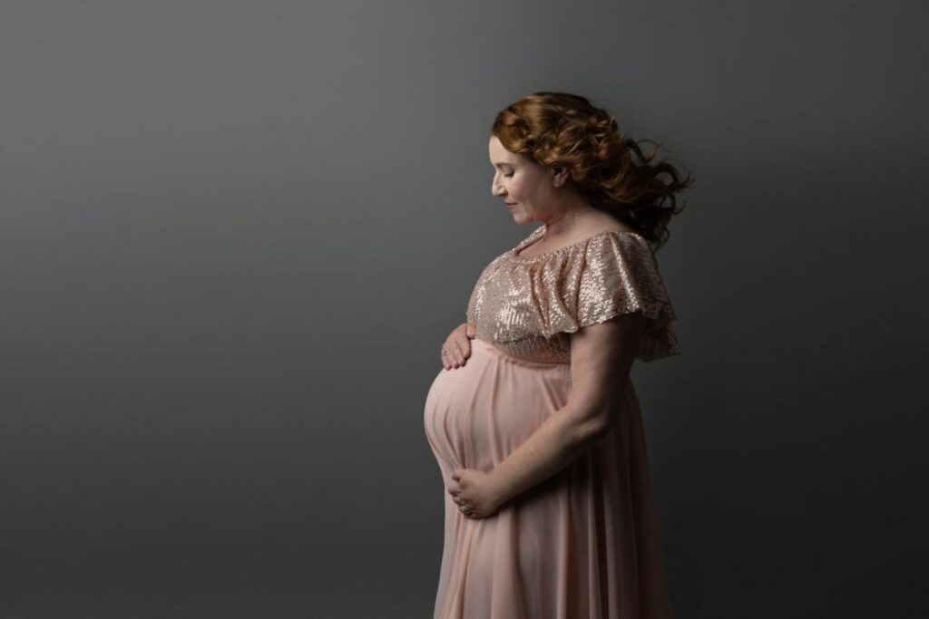 Woman wearing a long peach dress with a shimmery top, while cupping her pregnant belly for her maternity shoot
