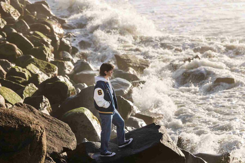 A student posing and standing on the big rocks with some waves crashing in front of him