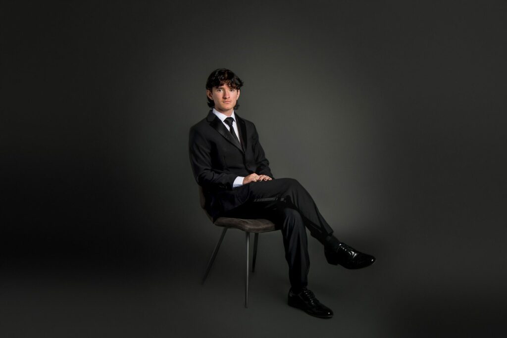 A student wearing a formal suit while sitting on a chair for his senior photograph