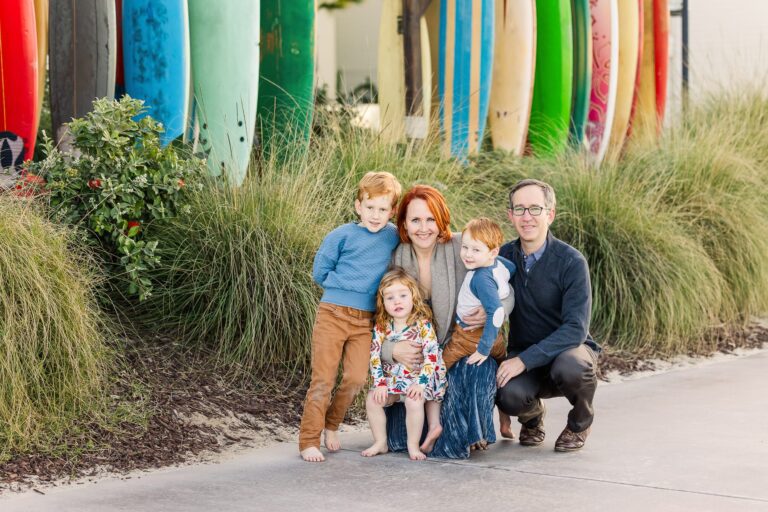 A family smiles together for a photo, standing in front of colorful surfboards on a sunny beach
