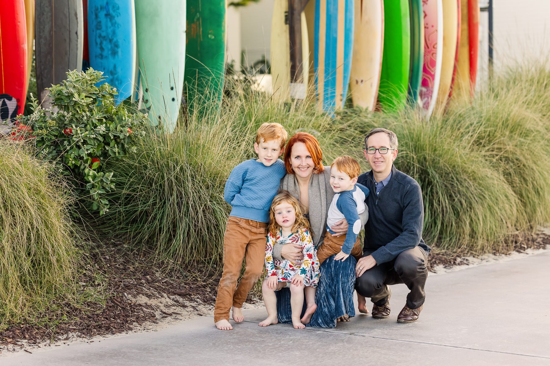 A family smiles together for a photo, standing in front of colorful surfboards on a sunny beach