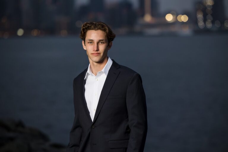 A young man in a suit standing in front of a city skyline with soft lighting