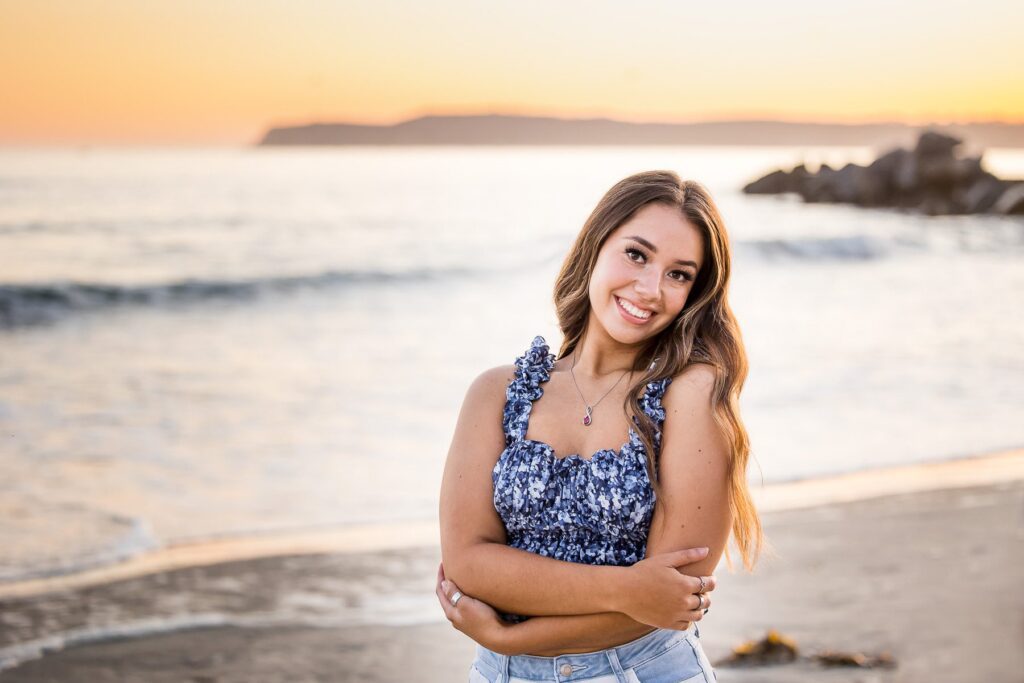 Young woman posing by the beach at sunset, smiling in a blue top