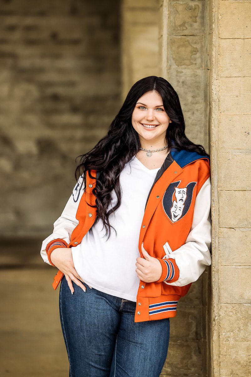 Smiling woman in a varsity jacket, leaning against a stone wall