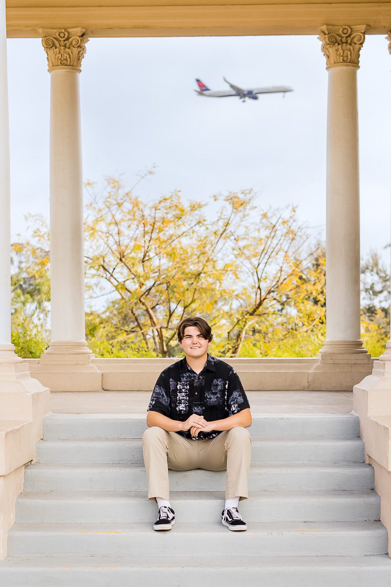 A young boy sits on steps, looking up at an airplane flying overhead against a clear blue sky