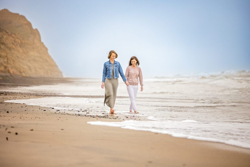 Two women walking barefoot on a beach near the shoreline