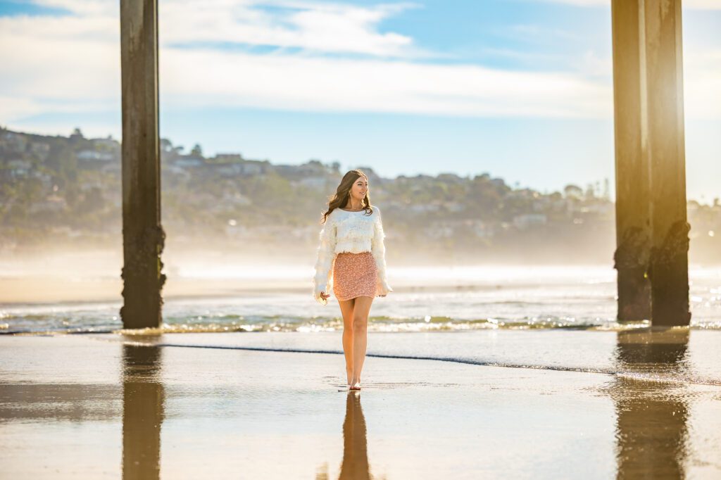 A young woman walks on the beach with a blue sky behind her and water below her.  She is walking underneath a cement pier and the light is warm and bright. 